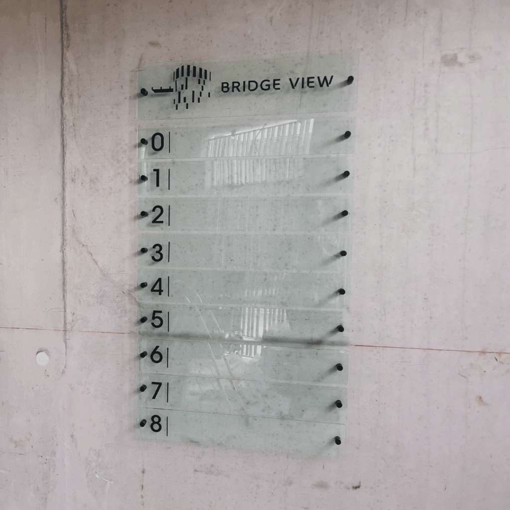 Signalétique directionnelle d'étages en verre givré pour le bâtiment Bridge View, indiquant les niveaux de 0 à 8 sur un mur en béton.