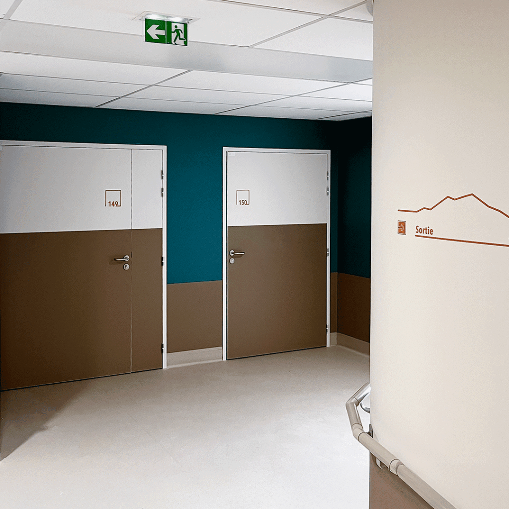 Couloir d'hôpital avec portes de chambres et signalétique murale d'orientation vers la sortie.