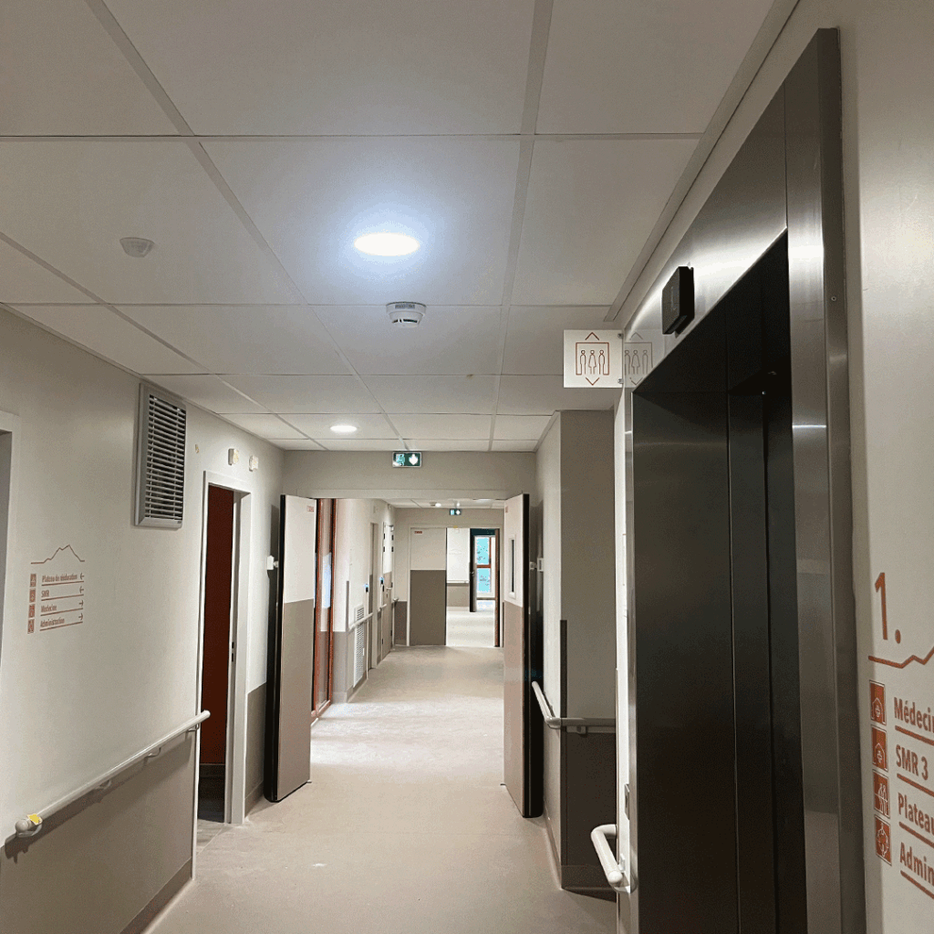 Couloir d'hôpital moderne avec portes colorées, ascenseur, et signalétique murale indiquant les services (Médecine - Hôpital de jour).