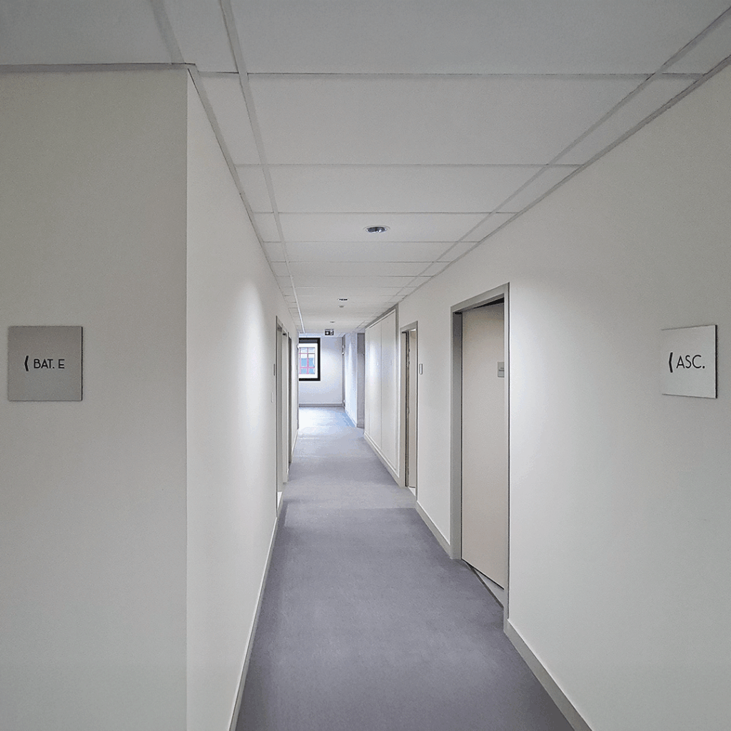 Plaques de signalétique carrées minimalistes fixées sur les murs blancs d'un long couloir de bureaux.