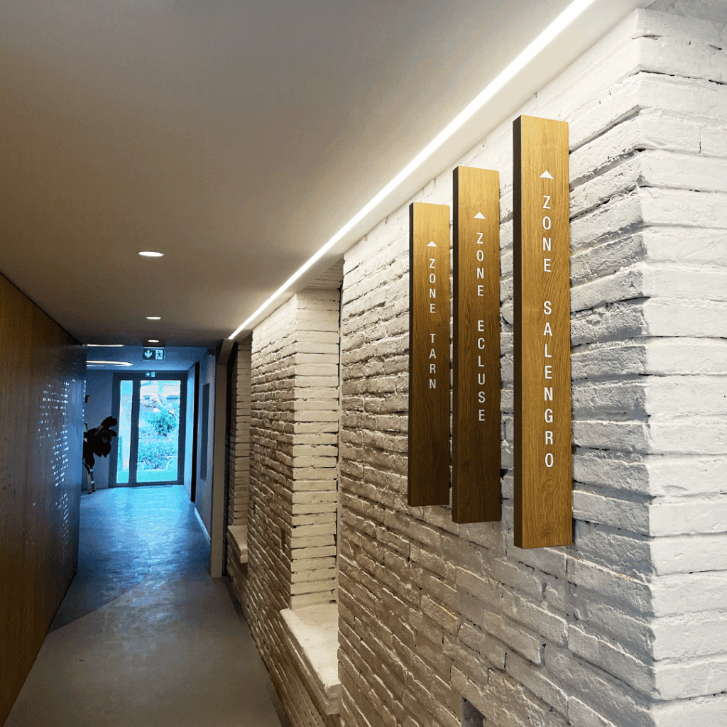 Trois panneaux de signalétique verticaux en bois brut avec inscriptions blanches, fixés sur un mur intérieur en briques.