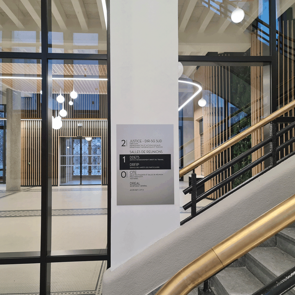 Plaque de signalétique intérieure sur poteau, indiquant les services (Justice, DREAL) à côté d'un escalier.