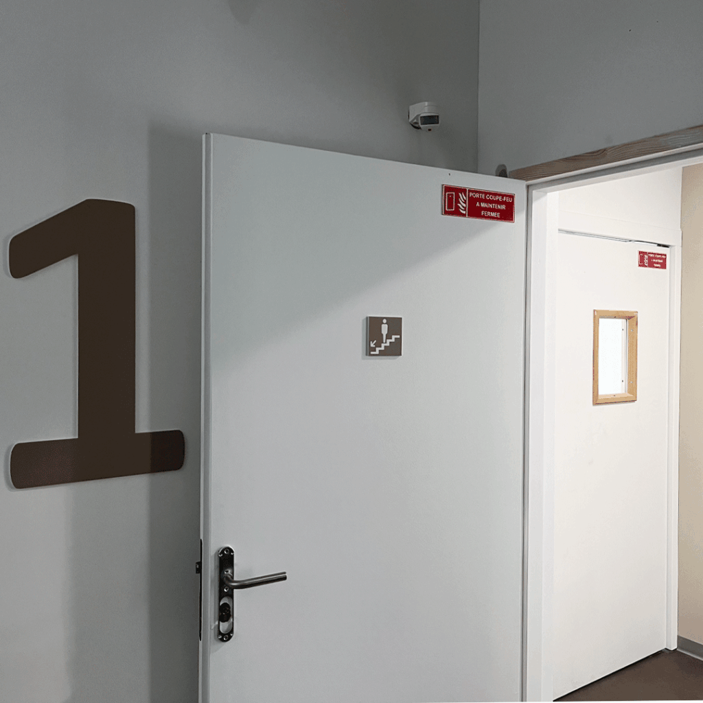 Signalétique d'étage en grand adhésif brun sur mur blanc près d'une porte d'accès identifiée avec une plaque de salle.