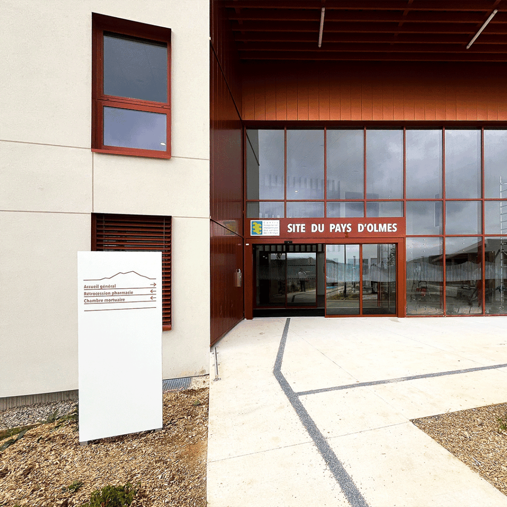 Totem blanc de signalétique extérieure à l'entrée d'un bâtiment moderne d'hôpital, indiquant l'accueil, la pharmacie et la chambre mortuaire.